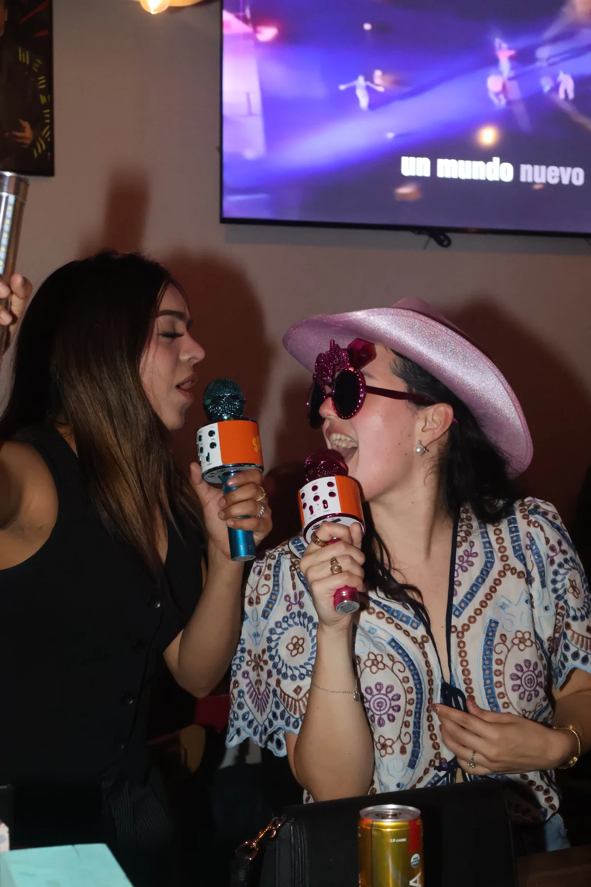 Two people sing into microphones beside a table; one wears a pink hat.