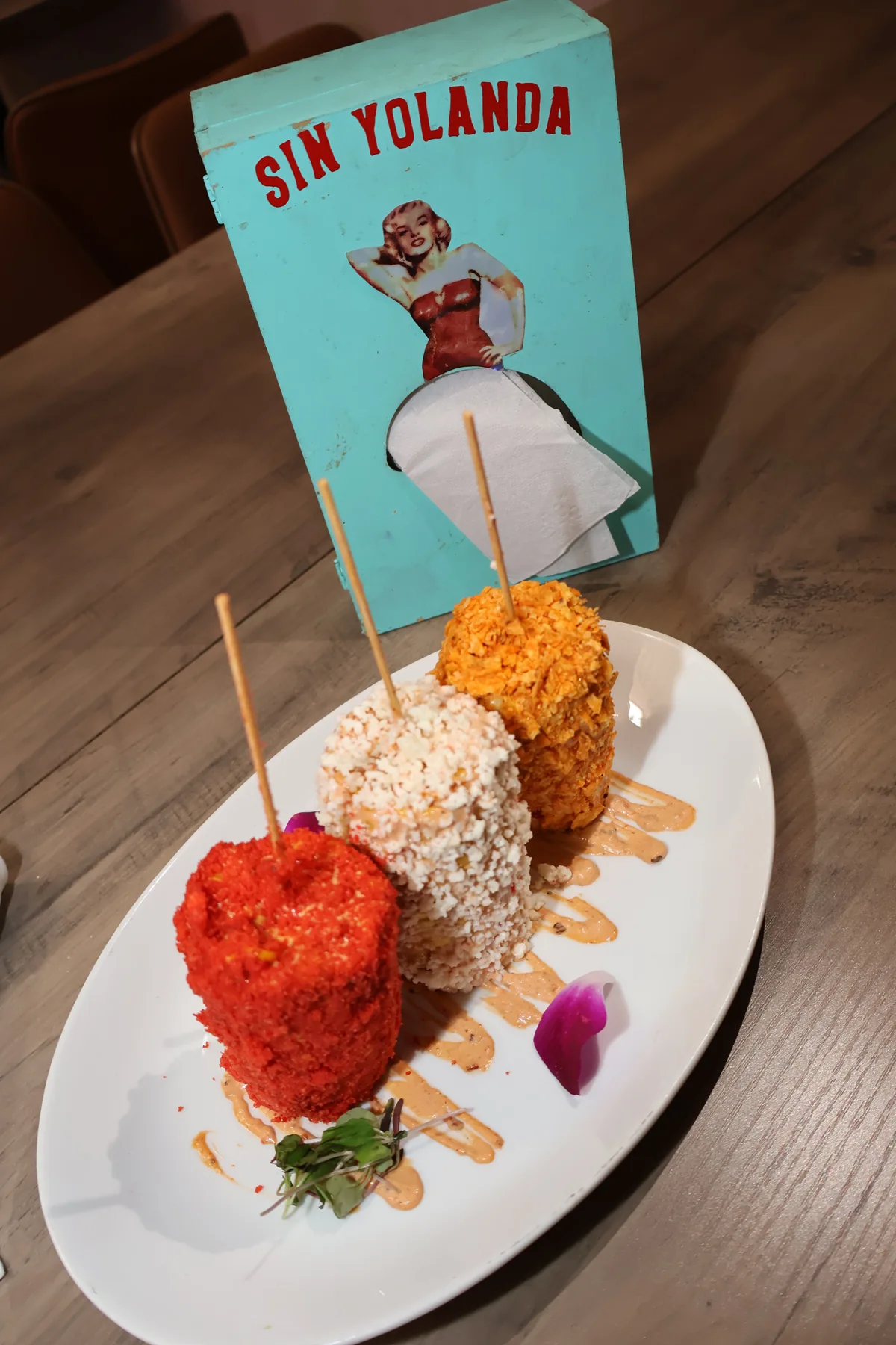 Three skewered bites with colorful toppings on a plate, with a Sin Yolanda napkin holder in the background.
