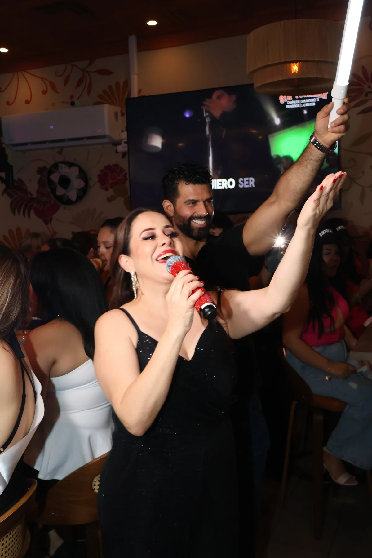 A person sings into a red microphone with an arm raised beside a group.