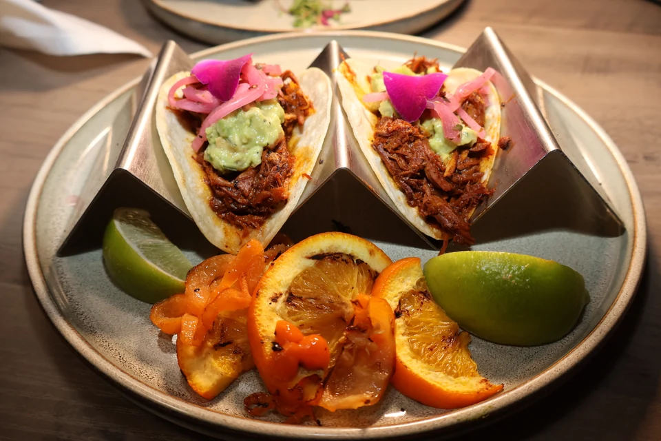 Two tacos on metal stands with citrus and a side dish on a gray plate.