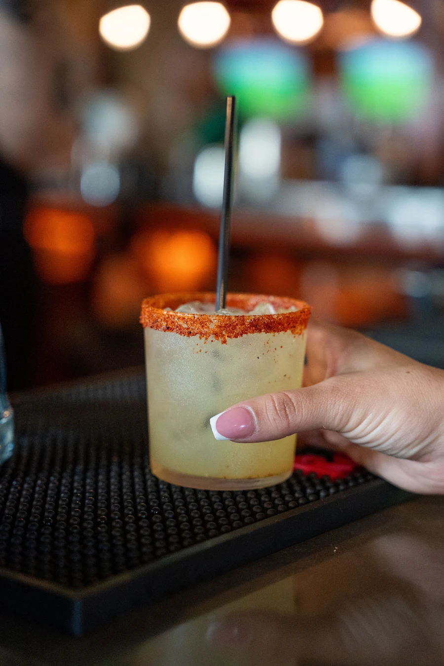 A hand holds a cocktail with a chili rim in front of the softly lit bar.